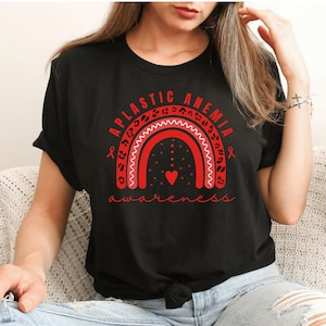 Può includere: T-shirt nera con un design arcobaleno rosso e leopardato e le parole "Aplastic Anemia Awareness". Il design include un cuore e nastri rossi di sensibilizzazione. La maglietta ha un girocollo.