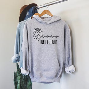 May include: A light gray hoodie with a black anatomical heart graphic and a heartbeat line. The text "DON'T BE TACHY" is printed below the graphic. The hoodie has a front pocket and a hood.