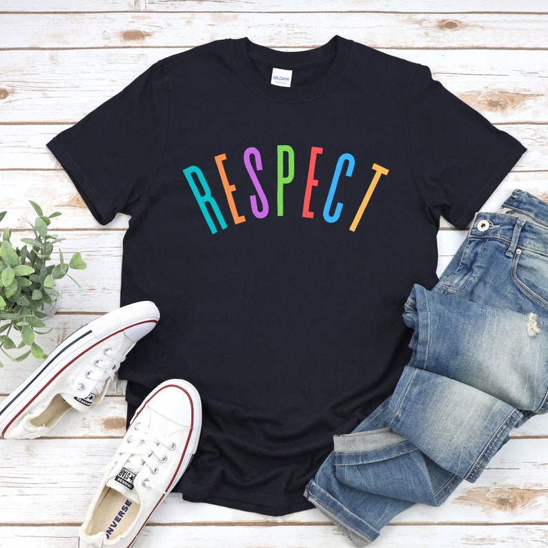 May include: A black t-shirt with the word "RESPECT" printed in rainbow colors on the front. The shirt is laid flat on a wooden surface with a pair of white sneakers and blue jeans.