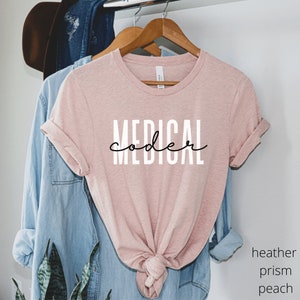 Pode incluir: Uma camiseta rosa com o texto "MEDICAL coder" em letras pretas e brancas.