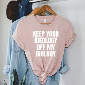 May include: A pink t-shirt with the text "Keep your ideology off my biology" printed in white.