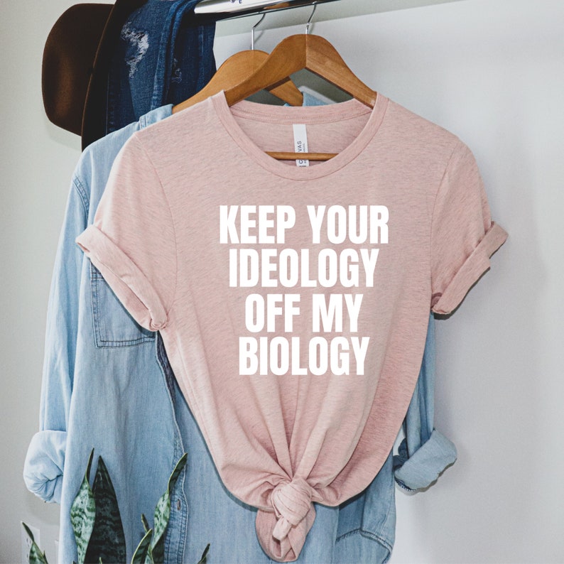 May include: A pink t-shirt with the text "Keep your ideology off my biology" printed in white.