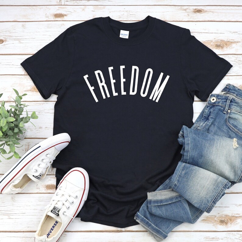 Op de afbeelding: Een zwart T-shirt met het woord "FREEDOM" in wit op de voorkant. Het shirt ligt op een wit houten oppervlak met een paar witte sneakers en een blauwe jeans.