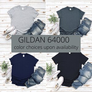 Op de afbeelding: Vier effen T-shirts in verschillende kleuren: grijs, donkerblauw, marineblauw en zwart. De T-shirts liggen op een witte houten achtergrond met jeans en sneakers. De tekst "GILDAN 64000" en "kleurkeuze afhankelijk van beschikbaarheid" staat onder de shirts.