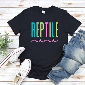 May include: Black t-shirt with the text "REPTILE mama" in rainbow colors. The shirt is folded and laid on a wooden surface with a pair of white sneakers and blue jeans.