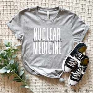 Könnte beinhalten: Ein graues T-Shirt mit dem Aufdruck "NUCLEAR technologist MEDICINE" in weiß. Das Shirt ist auf einem weißen, flauschigen Untergrund mit einem Paar schwarzen Converse-Turnschuhen zu sehen.