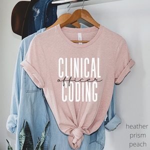 May include: A pink t-shirt with the text "CLINICAL officer CODING" in black and white lettering. The shirt is hanging on a wooden hanger.