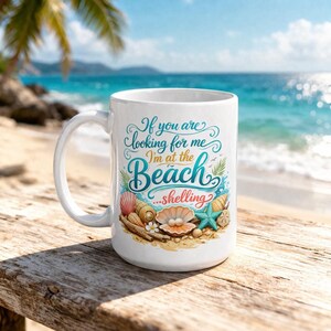 Puede incluir: Taza de cerámica blanca con un diseño de playa. La taza presenta el texto "If you are looking for me I'm at the Beach... shelling" con ilustraciones de conchas marinas, estrellas de mar y una perla. La taza está sobre una superficie de madera con un fondo de playa.