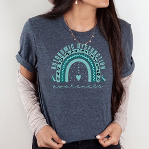 Puede incluir: Una camiseta azul turquesa con un gráfico de arcoíris que dice "Autonomic Dysfunction Awareness" con un pequeño corazón en el centro del arcoíris.
