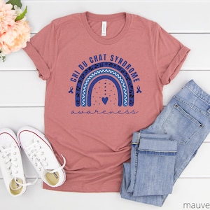 May include: Mauve t-shirt with a blue rainbow and the text "Cri du Chat Syndrome Awareness" in navy blue. The rainbow has a small heart in the center and two blue ribbons on either side.