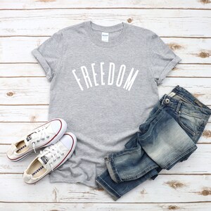 Op de afbeelding: Een grijs T-shirt met het woord "FREEDOM" in wit op de voorkant. Het shirt wordt gecombineerd met een blauwe jeans en witte sneakers.