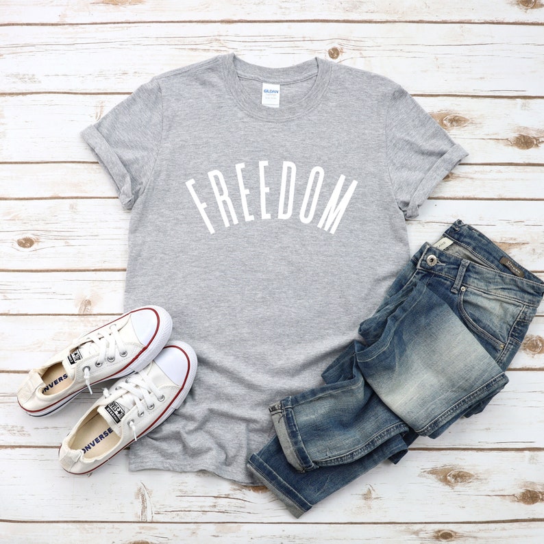 Op de afbeelding: Een grijs T-shirt met het woord "FREEDOM" in wit op de voorkant. Het shirt wordt gecombineerd met een blauwe jeans en witte sneakers.