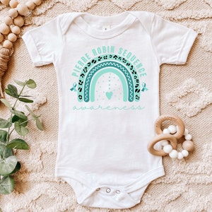 May include: White baby onesie with a turquoise rainbow design and the text "Pierre Robin Sequence Awareness". The rainbow has black leopard print spots and turquoise ribbons on each side.