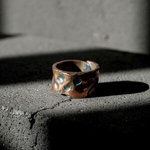 Peut inclure: Une large bague en cuivre présentant une surface martelée et irrégulière avec des zones en creux arborant une patine bleu sarcelle foncé. L'anneau possède un design ouvert et superposé avec un espace visible sur un côté, offrant un aspect métallique.
