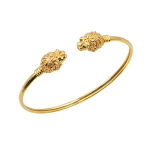 Op de afbeelding: Een goudkleurige manchetarmband met leeuwenkopdetails. De leeuwenkoppen bevinden zich aan elk uiteinde van de armband en hebben groene edelsteenogen. De armband is gemaakt van glanzend, glad metaal.