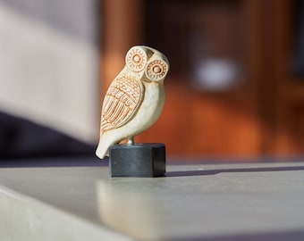 Puede incluir: Una pequeña figura de búho de color crema con detalles de líneas marrones grabadas en las alas y ojos circulares. El búho está montado sobre una base cuadrada negra sólida, creando una pieza decorativa estable con un acabado mate.