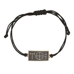 May include: A black cord bracelet with a silver rectangular charm featuring the text "T'O'" and a circle with a cross inside.