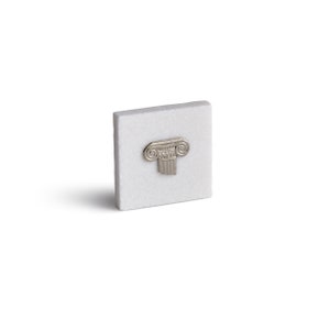 May include: A small white square tile with a silver metal column capital design.