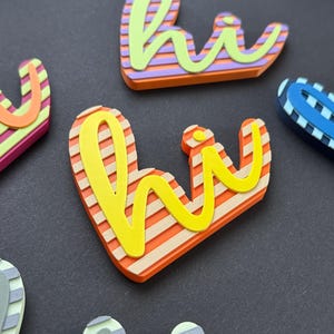 Könnte beinhalten: Nahaufnahme von farbenfrohen "hi" Wortkunst-Dekorationen. Die Buchstaben sind gelb, orange, rosa und blau, mit gestreiften und einfarbigen Mustern. Wahrscheinlich handelt es sich um Dekorationsartikel oder Verzierungen.