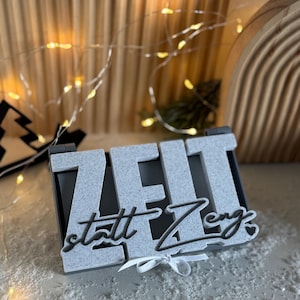 Op de afbeelding: Een decoratief bord met het woord "ZEIT" in grote, getextureerde, grijze letters en de zin "statt Zeng" in een scriptlettertype. Onderaan is een kleine witte strik vastgebonden. Het bord staat tegen een vage achtergrond met feeërieke lichtjes.