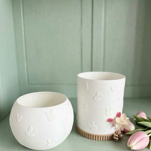Puede incluir: Dos jarrones de cerámica blanca con diseños en relieve de conejos y corazones. Uno es redondo, el otro cilíndrico con base acanalada. Tulipanes rosas y pequeñas flores se colocan cerca del jarrón cilíndrico.