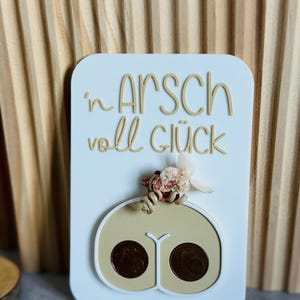 以下が含まれることがあります： ドイツ語のフレーズ「'n Arsch voll Glück」がベージュの文字で書かれた白い長方形のプラーク。プラークには、2つのコインと小さな花の配置が施された、ベージュ色の尻のデザインが施されています。