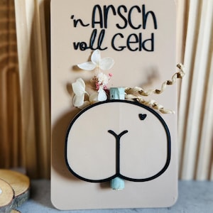 Puede incluir: Una tarjeta beige con la frase alemana "'n Arsch voll Geld" en letras negras, con un dise&ntilde;o de trasero de dibujos animados con un contorno negro. La tarjeta est&aacute; decorada con flores secas y un peque&ntilde;o coraz&oacute;n.
