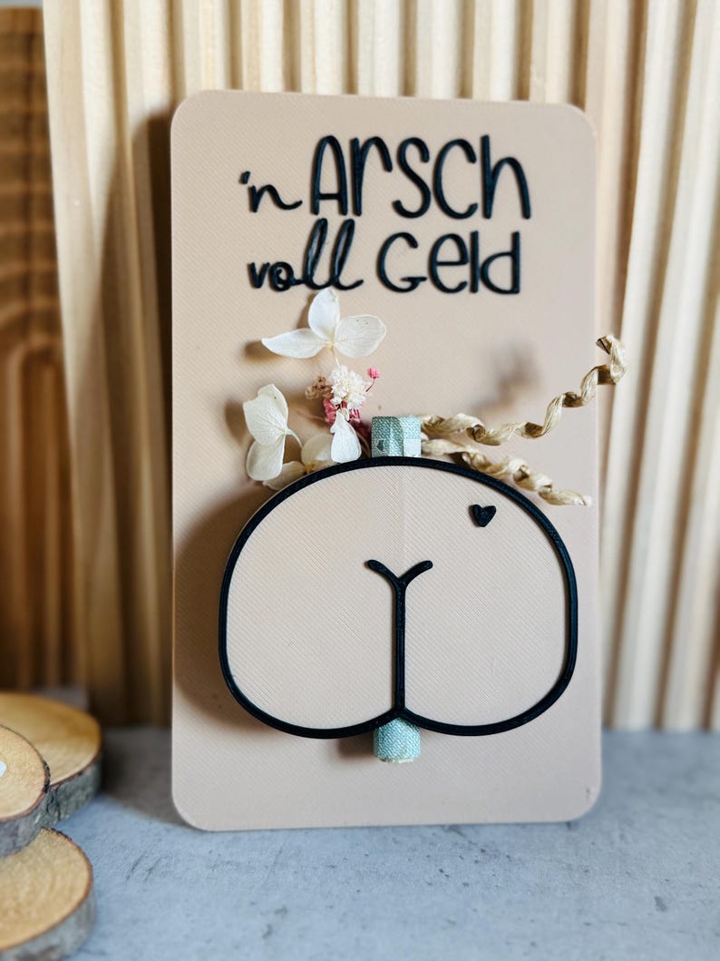 Puede incluir: Una tarjeta beige con la frase alemana "'n Arsch voll Geld" en letras negras, con un dise&ntilde;o de trasero de dibujos animados con un contorno negro. La tarjeta est&aacute; decorada con flores secas y un peque&ntilde;o coraz&oacute;n.