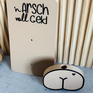 Puede incluir: Un letrero rectangular beige con el texto "'n ARSCH voll GELD" en escritura negra. Junto a &eacute;l, un objeto ovalado beige con contorno negro y un detalle de coraz&oacute;n. Los objetos est&aacute;n sobre una superficie gris claro.