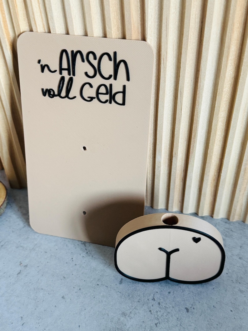 Puede incluir: Un letrero rectangular beige con el texto "'n ARSCH voll GELD" en escritura negra. Junto a &eacute;l, un objeto ovalado beige con contorno negro y un detalle de coraz&oacute;n. Los objetos est&aacute;n sobre una superficie gris claro.