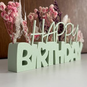Könnte beinhalten: Ein mintgrünes "HAPPY BIRTHAY"-Schild mit einem Herz, das das "O" in Geburtstag ersetzt. Das Schild enthält rosa und lila Trockenblumen. Der Hintergrund ist hellbraun.