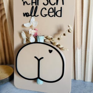 Puede incluir: Una tarjeta beige con una ilustraci&oacute;n de trasero de dibujos animados, un peque&ntilde;o coraz&oacute;n y el texto alem&aacute;n "in Arsch voll Geld" en negro. La tarjeta est&aacute; decorada con flores secas y una cinta.
