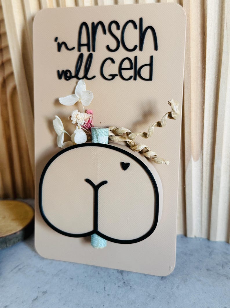 Puede incluir: Una tarjeta beige con una ilustraci&oacute;n de trasero de dibujos animados, un peque&ntilde;o coraz&oacute;n y el texto alem&aacute;n "in Arsch voll Geld" en negro. La tarjeta est&aacute; decorada con flores secas y una cinta.