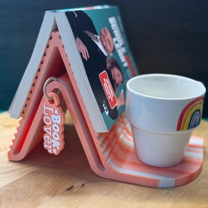 May include: A coral-coloured book holder with a matching coaster, holding a book. The holder has a layered design with the words "Book Lover" on a charm. A white mug with a rainbow design sits on the coaster.