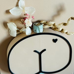 Puede incluir: Una tarjeta de color beige claro con un contorno negro de una cara de oso, un peque&ntilde;o coraz&oacute;n negro y las palabras alemanas "voll Geld" en negro. La tarjeta est&aacute; decorada con flores blancas secas, capullos rosas y un peque&ntilde;o regalo turquesa y dorado.