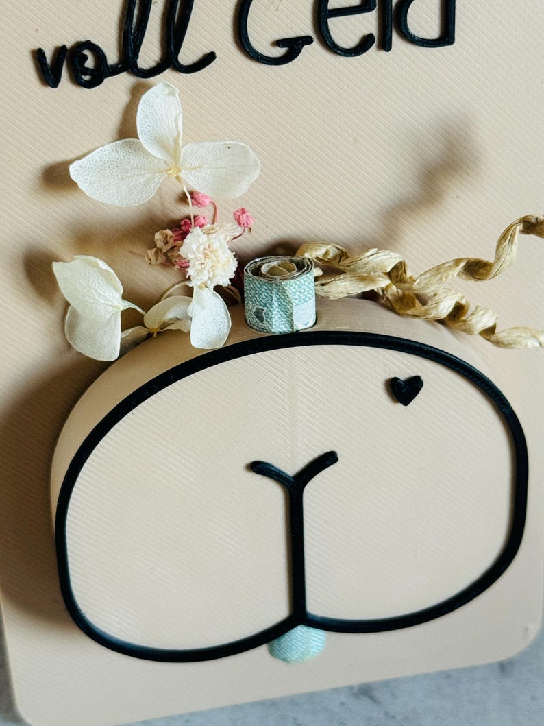 Puede incluir: Una tarjeta de color beige claro con un contorno negro de una cara de oso, un peque&ntilde;o coraz&oacute;n negro y las palabras alemanas "voll Geld" en negro. La tarjeta est&aacute; decorada con flores blancas secas, capullos rosas y un peque&ntilde;o regalo turquesa y dorado.