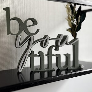Könnte beinhalten: Ein dekoratives Regal-Akzent mit den Worten "be you tiful" in einem geschichteten 3D-Design. Die Buchstaben sind ein gedämpftes Grün mit einer helleren grauen Kontur. Das Regal enthält auch getrocknete Blumen und Grün.