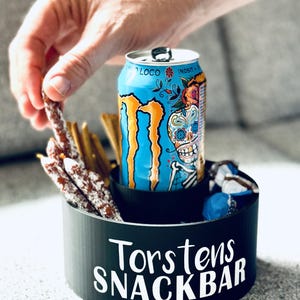 Könnte beinhalten: Ein schwarzer Snackhalter mit dem Text "Torstens SnackBar" in weißen Buchstaben. Eine Dose Monster-Energy-Drink mit einem Zucker Schädel-Design ist im Halter. Der Halter enthält auch eine Auswahl an Snacks, darunter Brezeln und Wurststäbchen.
