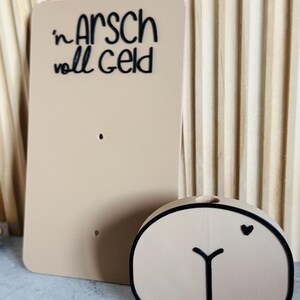 Puede incluir: Un letrero rectangular beige con la frase "in Arsch voll Geld" en escritura negra. Junto a &eacute;l, un objeto beige con forma de trasero con un contorno negro y un peque&ntilde;o coraz&oacute;n negro.