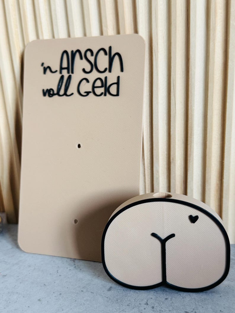 Puede incluir: Un letrero rectangular beige con la frase "in Arsch voll Geld" en escritura negra. Junto a &eacute;l, un objeto beige con forma de trasero con un contorno negro y un peque&ntilde;o coraz&oacute;n negro.