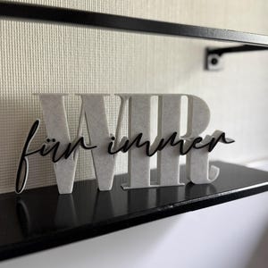 Pode incluir: Uma caneca de cerâmica branca com uma alça preta e o texto "But First, Coffee" em negrito preto. A caneca está sobre um fundo desfocado, sugerindo um ambiente acolhedor.