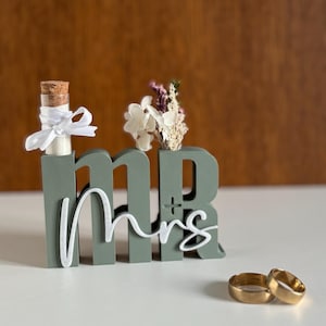 Könnte beinhalten: Eine salbeigrüne "Mr & Mrs" Hochzeitsdekoration mit einer kleinen Glasflasche und einem kleinen Blumenstrauß. Zwei goldene Eheringe befinden sich vor der Dekoration. Die Buchstaben sind in Blockform mit dem Wort "Mrs" in Schreibschrift.