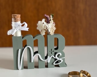May include: A sage green "Mr & Mrs" wedding decoration with a small glass bottle and a small bouquet of flowers. Two gold wedding bands are in front of the decoration. The letters are in a block style with the word "Mrs" in a cursive font.