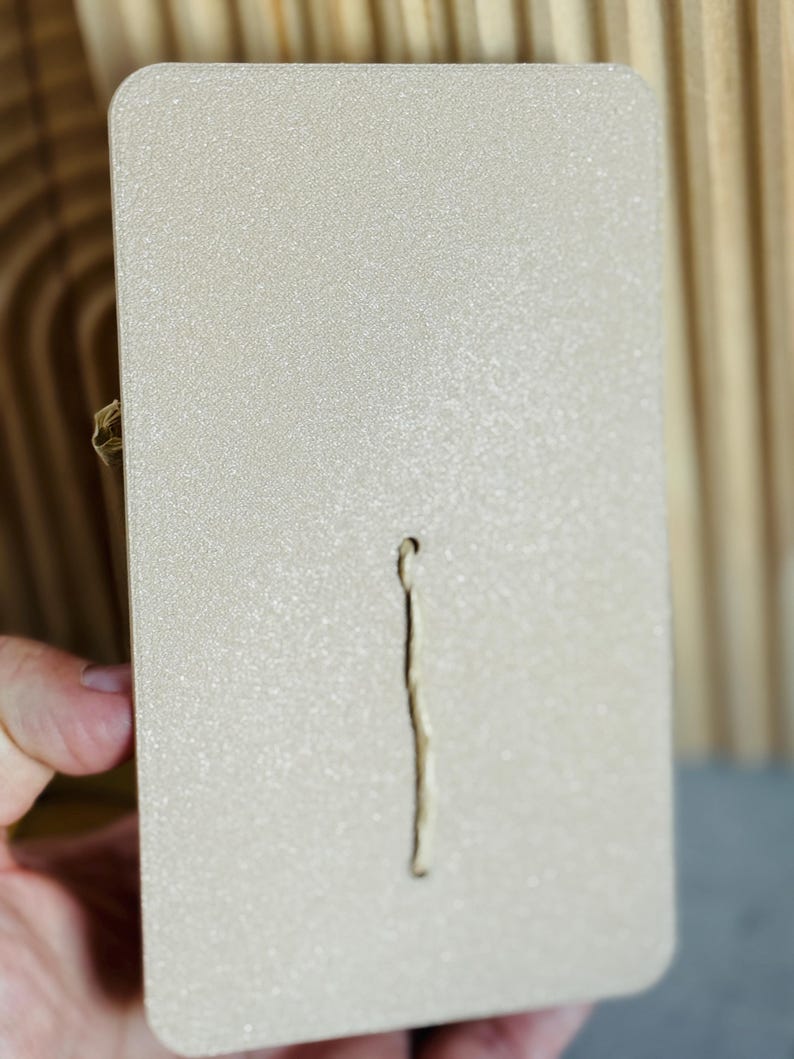 Puede incluir: Un art&iacute;culo rectangular de color beige claro con una superficie texturizada y una ranura vertical, estrecha y empotrada. El art&iacute;culo tiene esquinas redondeadas y un peque&ntilde;o cierre dorado en el lateral. El fondo es una superficie de madera marr&oacute;n claro.