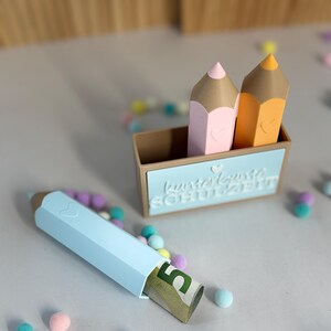 Könnte beinhalten: Ein brauner, bleistiftförmiger Behälter mit einem rosa und einem orangefarbenen Stift, jeweils mit einem kleinen Herzdetail. Ein separater hellblauer Stiftbehälter liegt davor und enthält einen gerollten Fünf-Euro-Schein. Kleine bunte Pompons.