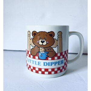May include: White ceramic mug with a cartoon bear design. The bear is in bed and holding a blue cup. The mug has a red and white checkered pattern and the words "LITTLE DIPPER" in blue.