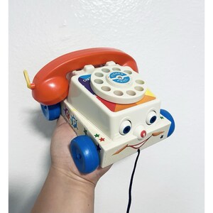 May include: Vintage Fisher-Price Chatter Telephone toy. The toy phone has a white body, orange handset, and blue wheels. The rotary dial is white with the Fisher-Price logo. The phone has a smiling face and a pull string.
