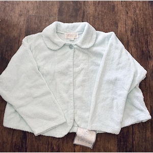May include: A light blue terry cloth jacket with a rounded collar and a single button closure. The jacket has long sleeves and a tag that reads "HERBCRAFT". The jacket is displayed on a wooden surface.