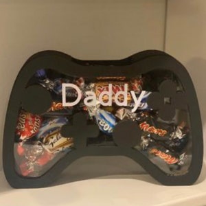 May include: A black, wooden, video game controller shaped box filled with assorted chocolate candy bars. The box has a white inscription that reads "Daddy".