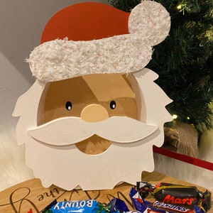 May include: A festive wooden Santa Claus face with a red hat and white beard, designed to hold sweets. The hat trim is white and sparkly. Assorted chocolate bars are arranged in front on a wooden tray.